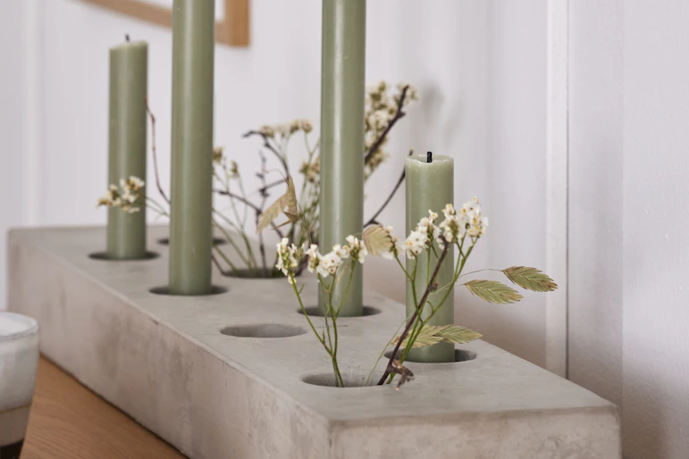 En ljusstake i grå cement har gröna kronljus och blommor i sig.