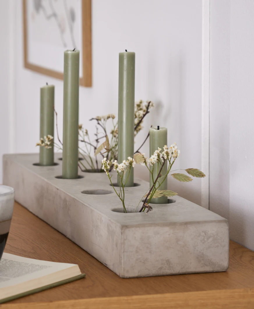 En ljusstake i grå cement har gröna kronljus och blommor i sig.