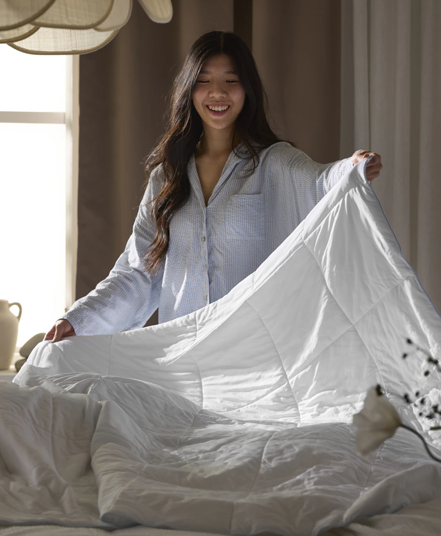 En brunhårig tjej i blå pyjamas håller i ett täcke och bäddar en säng