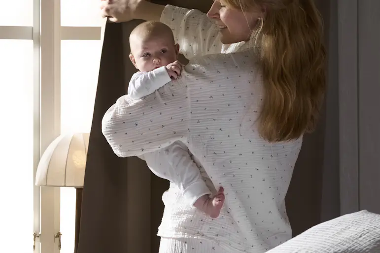 en mamma står i pyjamas vid ett fönster och håller i sin bebis