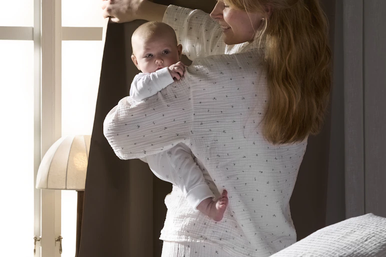 en mamma står i pyjamas vid ett fönster och håller i sin bebis