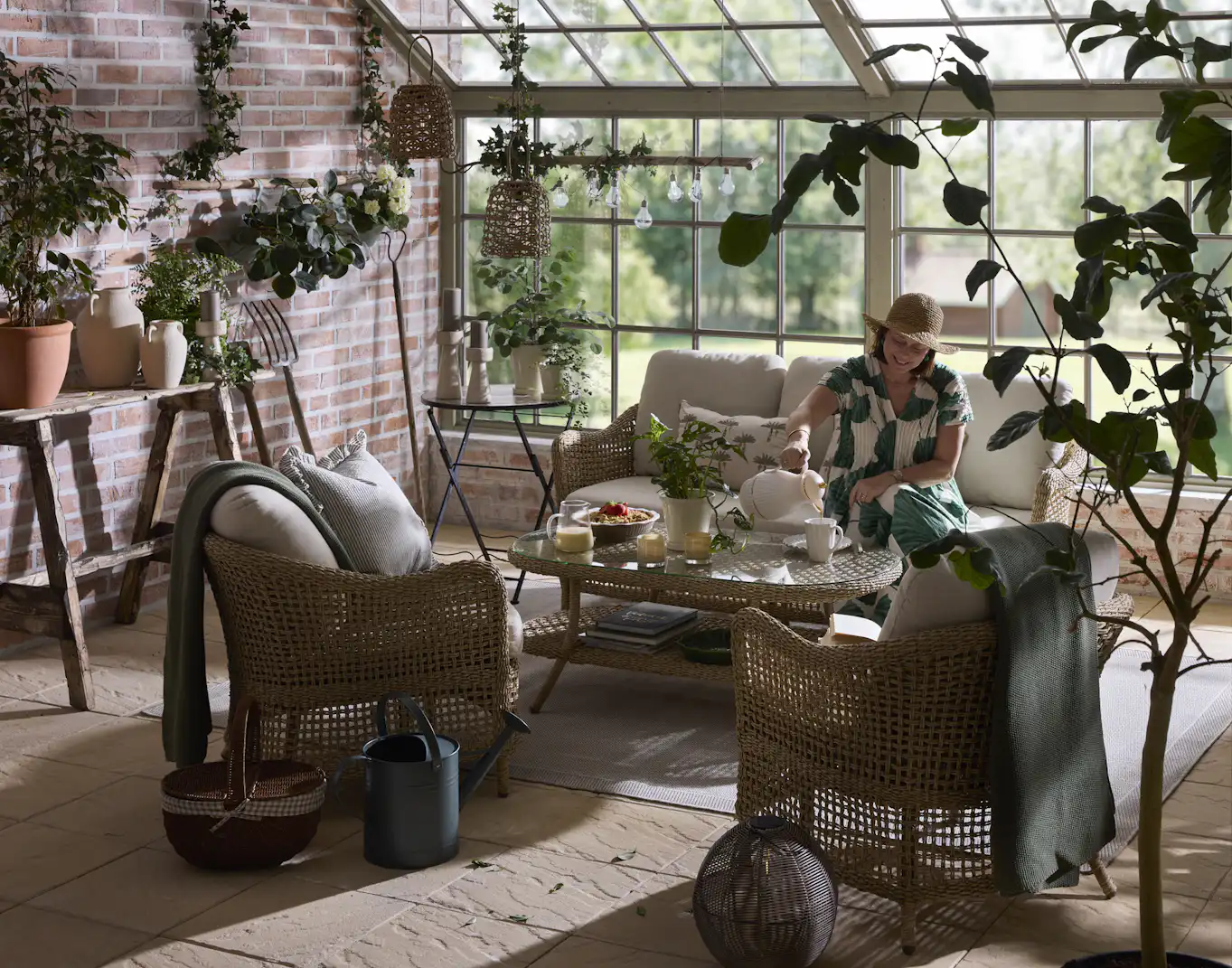 I ett orangerie sitter en kvinna med solhatt i en loungegrupp i konstrotting