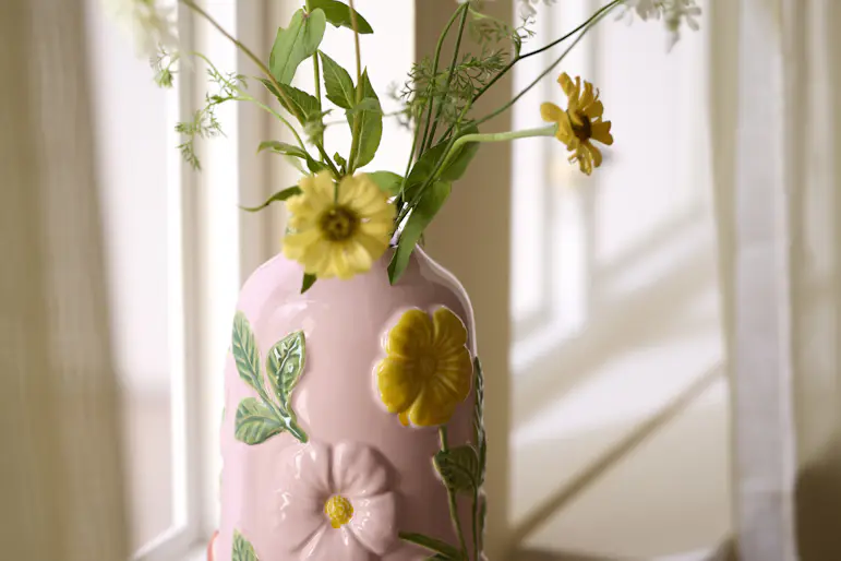 en rosa vas med blommor står i ett fönster med vita gardiner