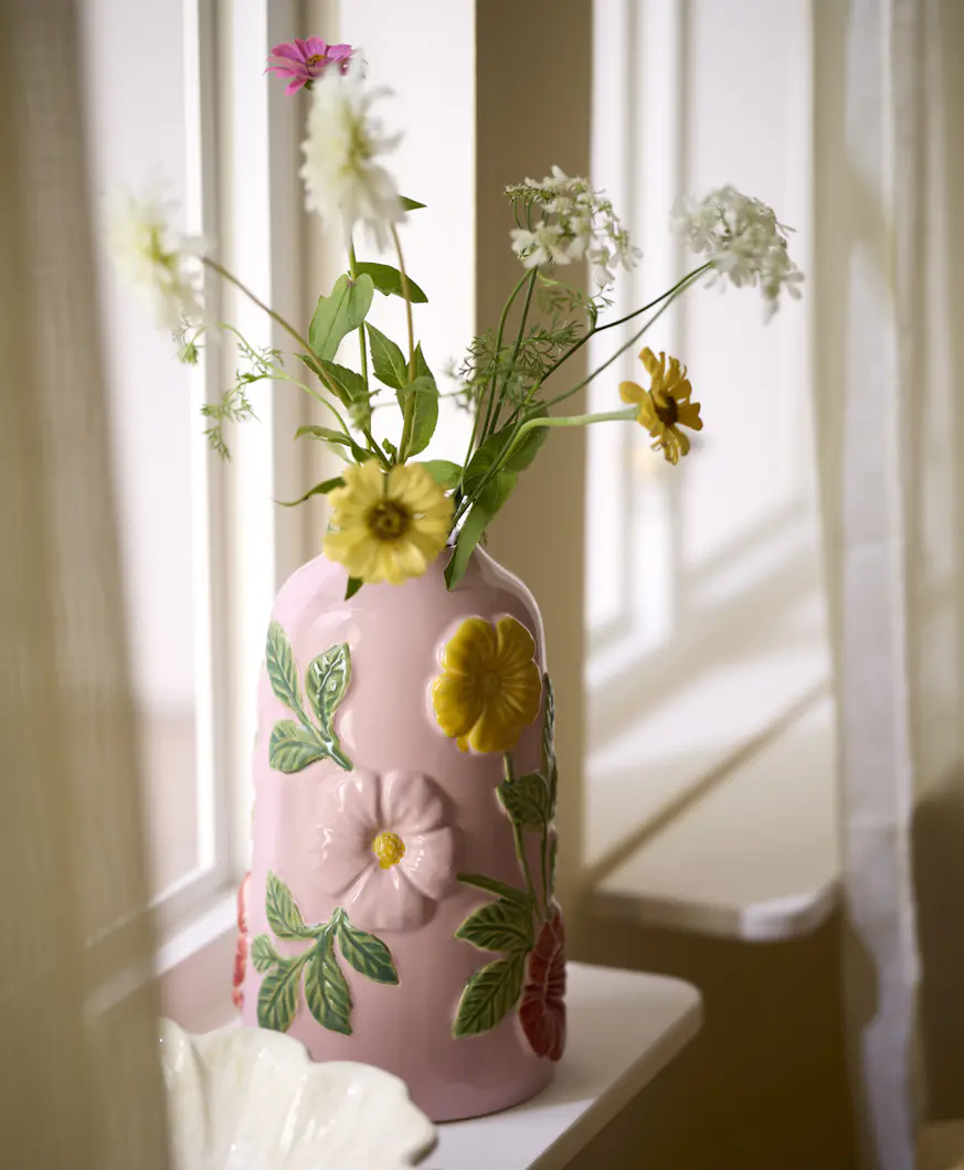 en rosa vas med blommor står i ett fönster med vita gardiner