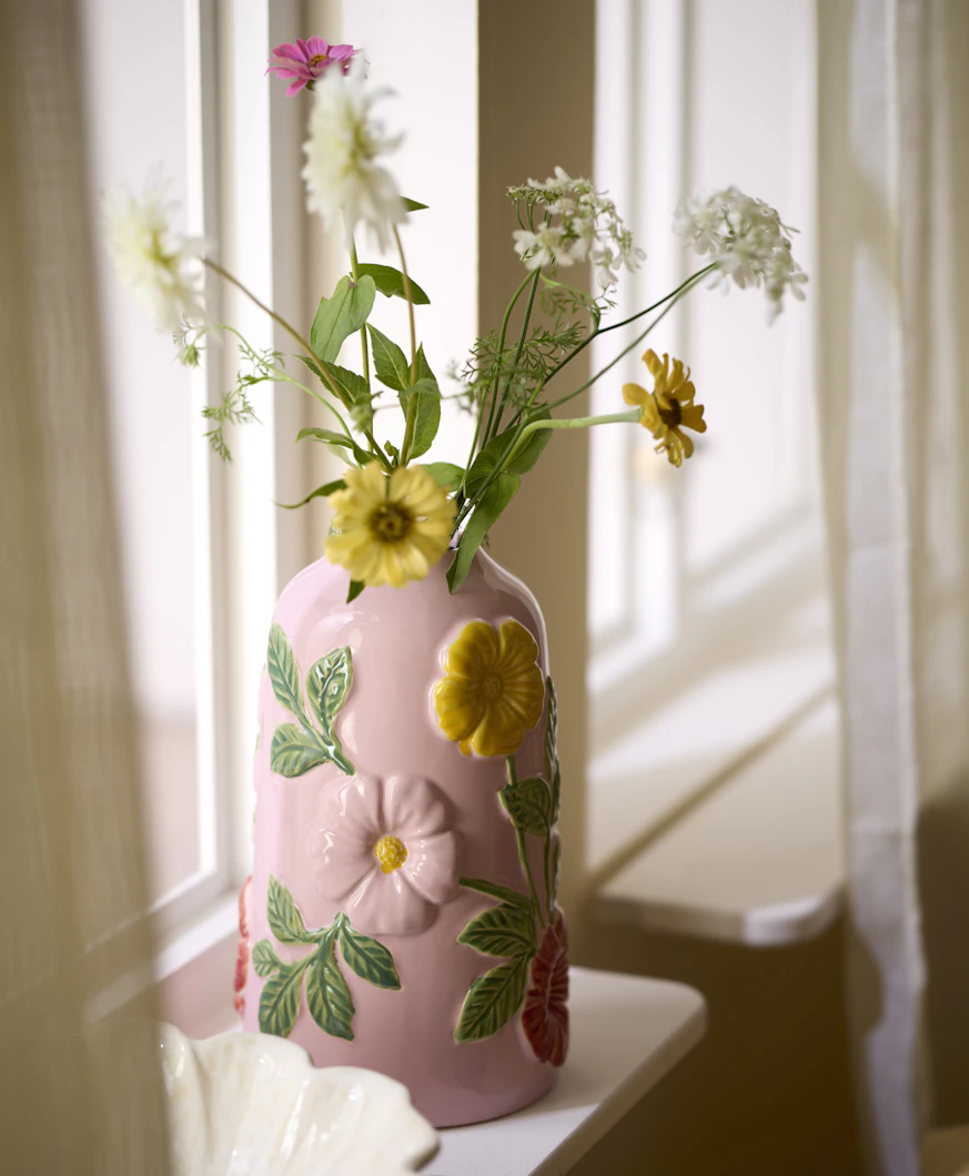 en rosa vas med blommor står i ett fönster med vita gardiner