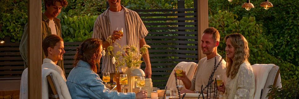 kvällsmiljö i skymning på en uteplats där en grupp människor sitter runt ett bord och pratar och skrattar.