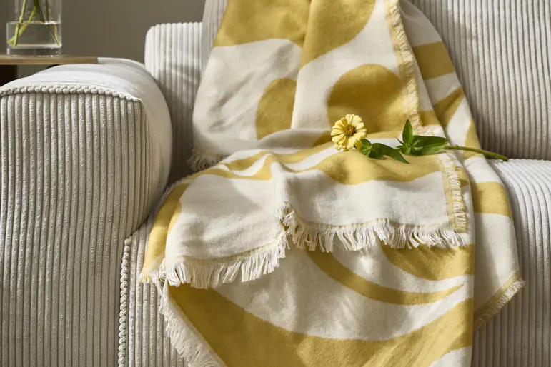 en gulmönstrad pläd ligger på kanten av en beige soffa tillsammans med gula konstväxter