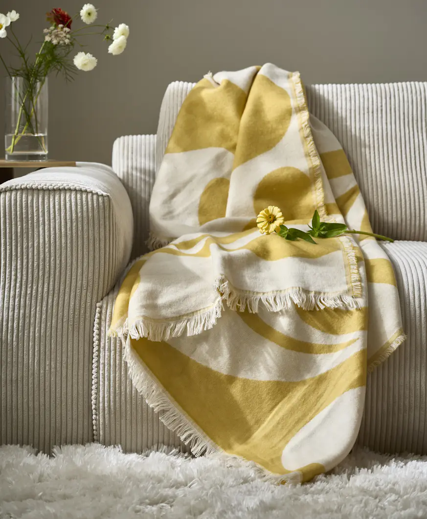 en gulmönstrad pläd ligger på kanten av en beige soffa tillsammans med gula konstväxter
