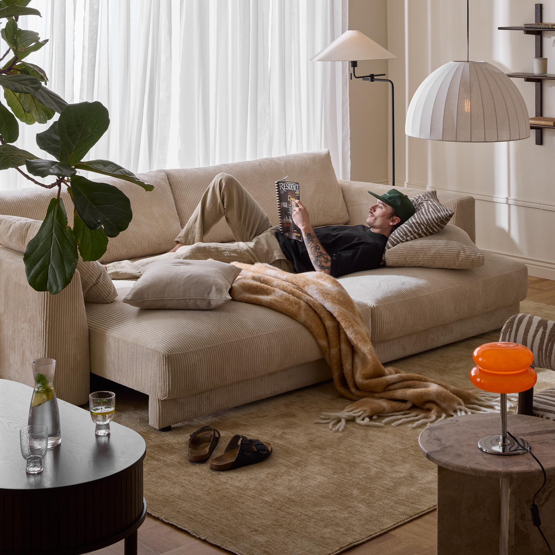 I ett vardagsrum står en bäddsoffa i vit manchester. I soffan ligger en ung man och läser