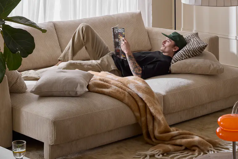 I ett vardagsrum står en bäddsoffa i vit manchester. I soffan ligger en ung man och läser