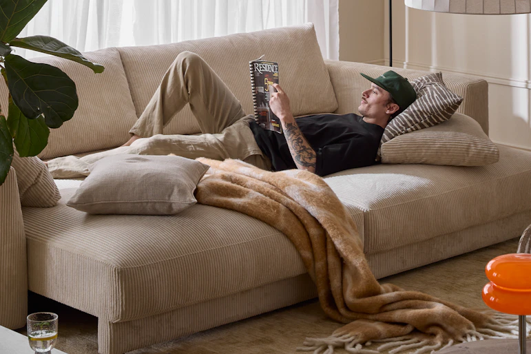 I ett vardagsrum står en bäddsoffa i vit manchester. I soffan ligger en ung man och läser