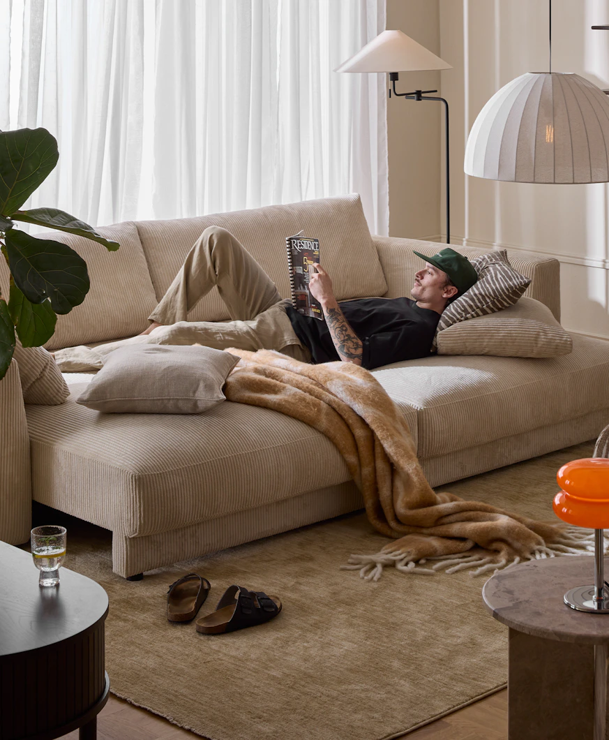 I ett vardagsrum står en bäddsoffa i vit manchester. I soffan ligger en ung man och läser