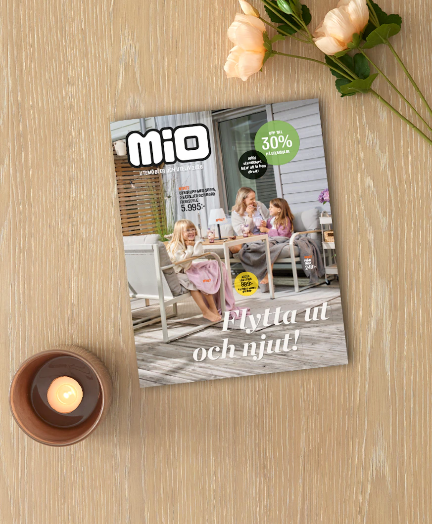 På ett ljust träbord ligger Mios utelivsbilaga intill ett ljus och en blomma.