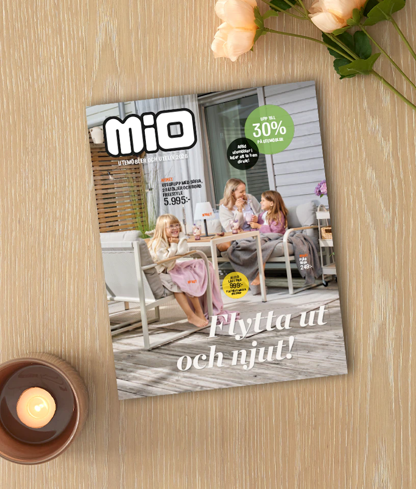 På ett ljust träbord ligger Mios utelivsbilaga intill ett ljus och en blomma.