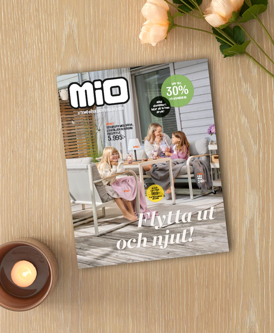 På ett ljust träbord ligger Mios utelivsbilaga intill ett ljus och en blomma.