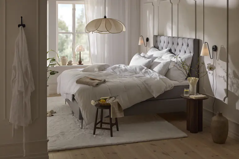 Ljust sovrum med bäddad säng i vita nyanser, solstrålar som kommer in från fönstret