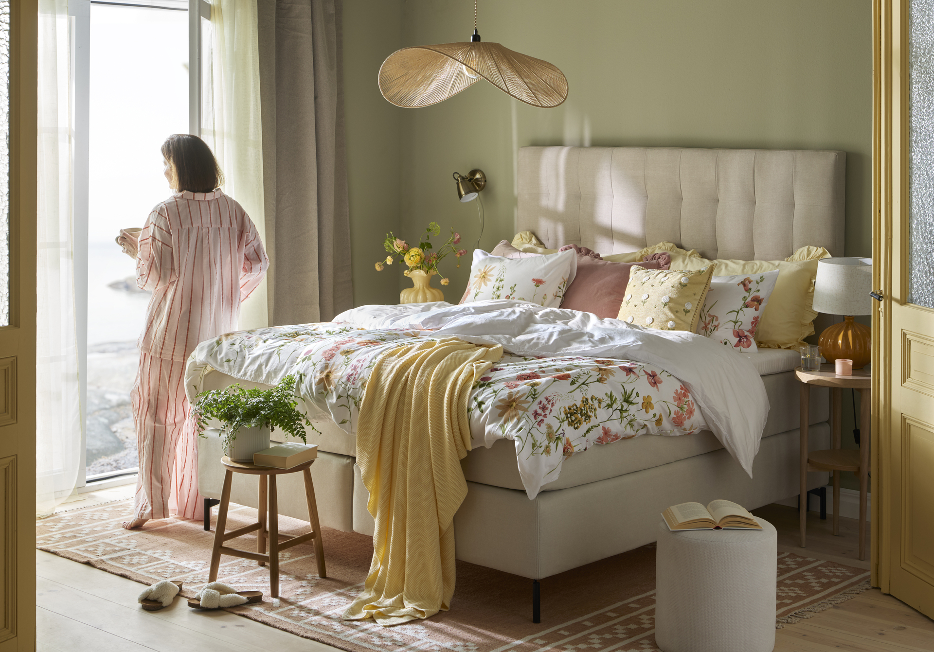 Säng med blommiga bäddtextilier, kvinna i pyjamas som står och tittar ut genom en öppen dörr.