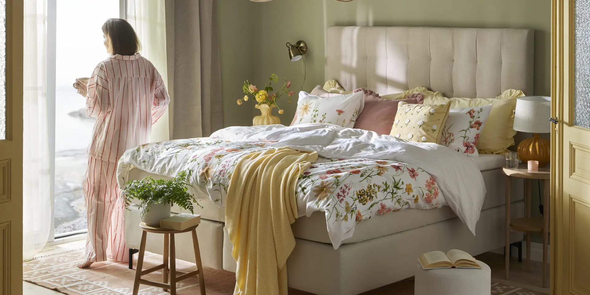 Säng med blommiga bäddtextilier, kvinna i pyjamas som står och tittar ut genom en öppen dörr.