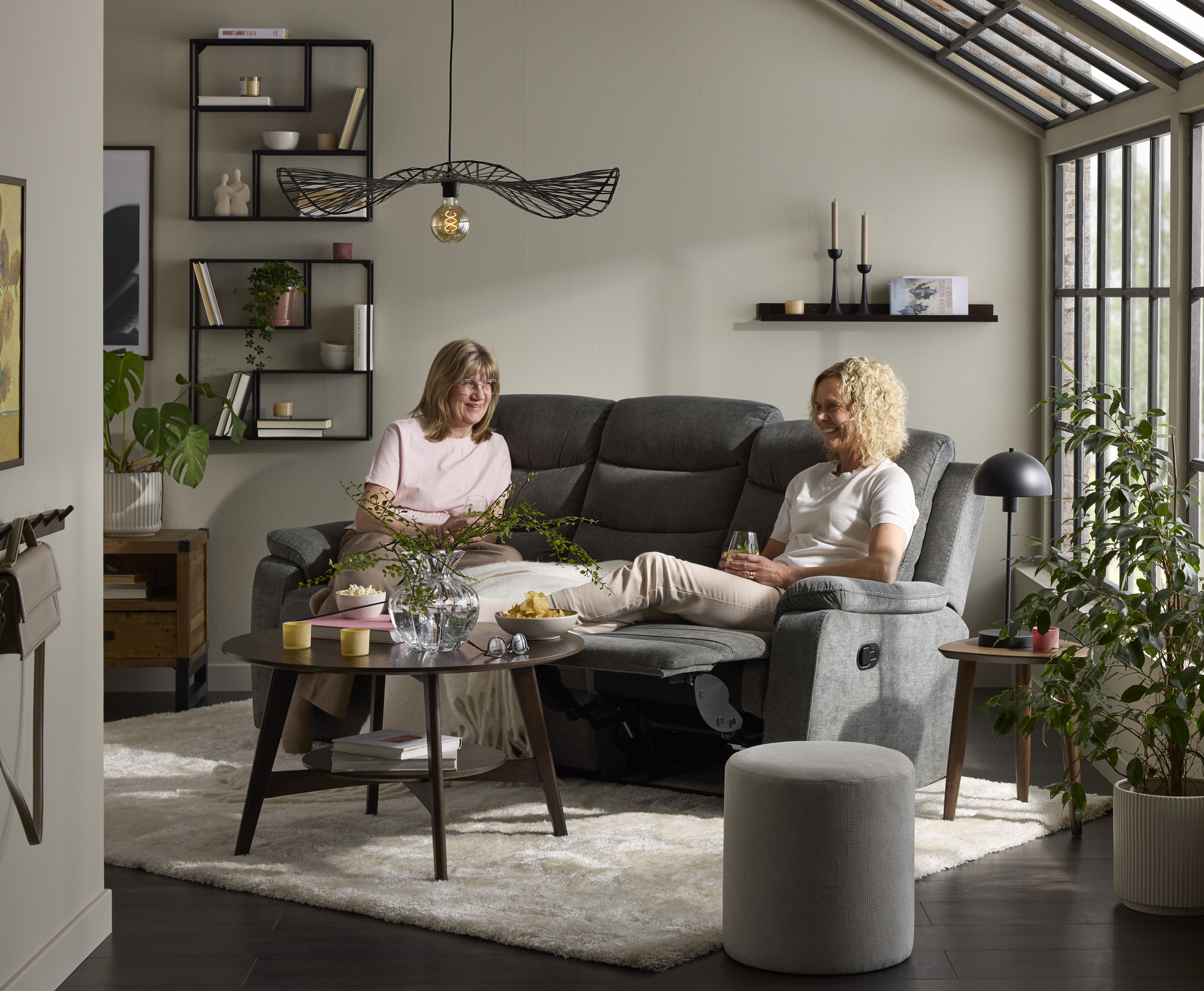  Två kvinnor sitter i en grå soffa med recliner i ett vardagsrum. Framför soffan står ett runt soffbord i mörkt trä.