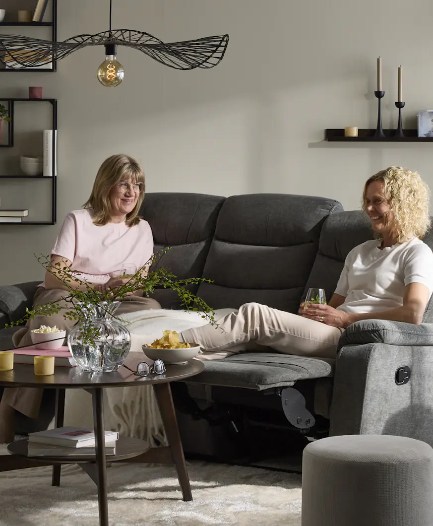 Två kvinnor sitter i en grå soffa med recliner i ett vardagsrum. Framför soffan står ett runt soffbord i mörkt trä.