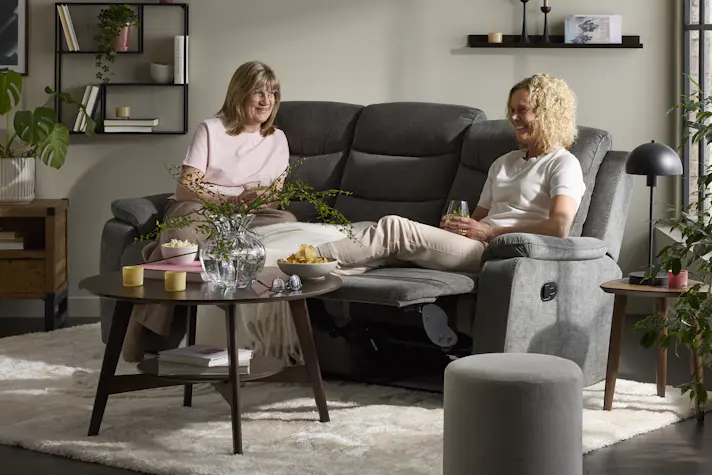 Två kvinnor sitter i en grå soffa med recliner i ett vardagsrum. Framför soffan står ett runt soffbord i mörkt trä.