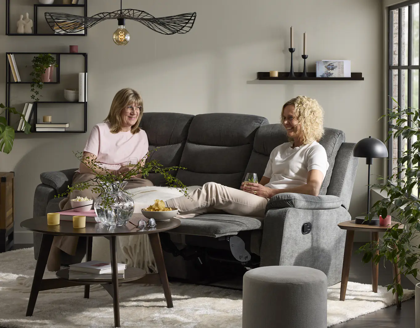Två kvinnor sitter i en grå soffa med recliner i ett vardagsrum. Framför soffan står ett runt soffbord i mörkt trä.