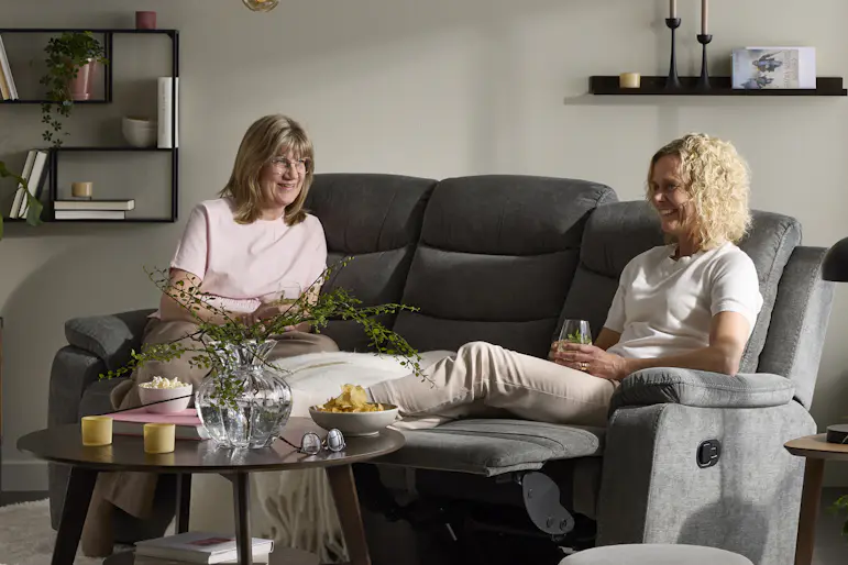 Två kvinnor sitter i en grå soffa med recliner i ett vardagsrum. Framför soffan står ett runt soffbord i mörkt trä.