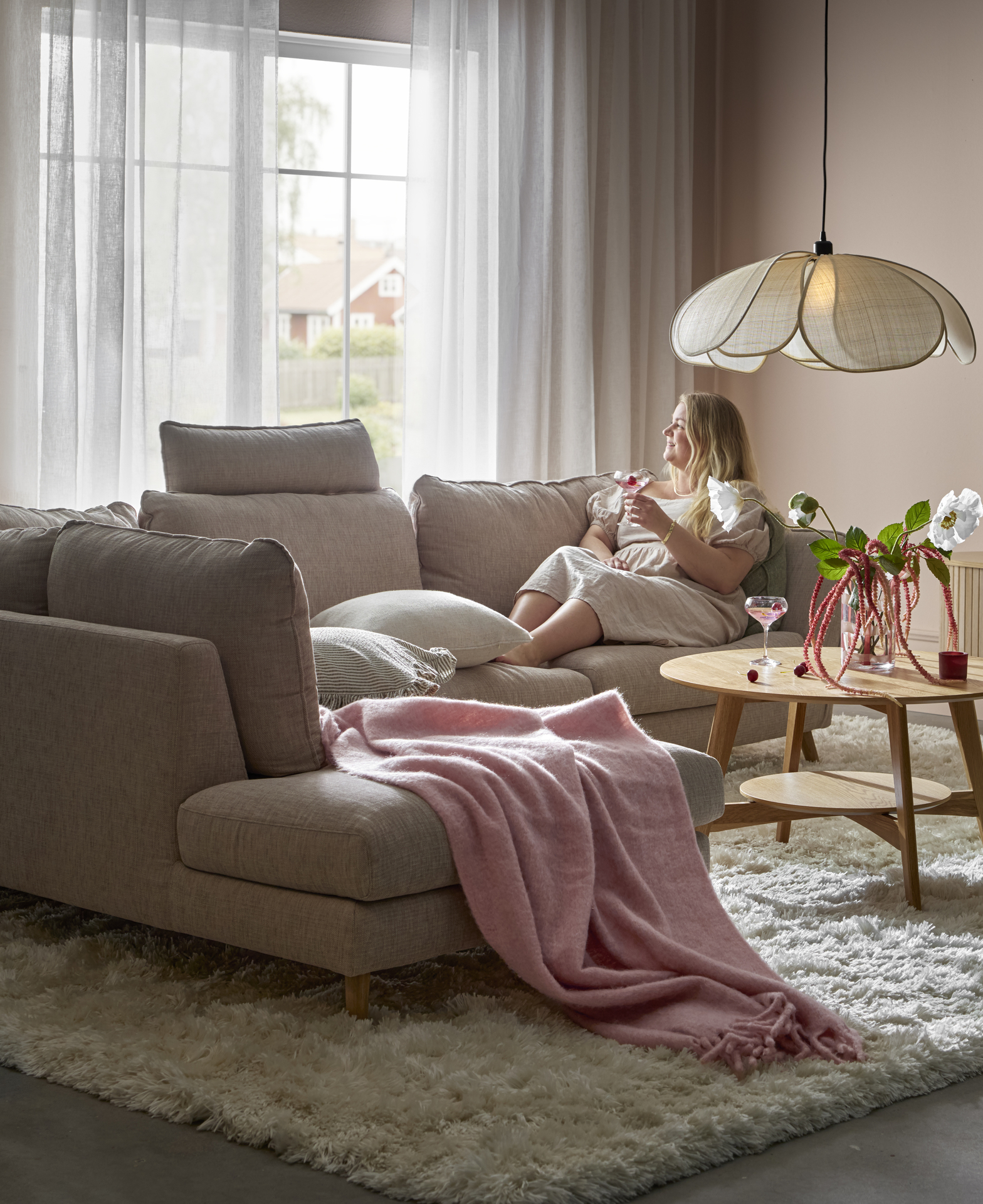 En kvinna sitter i en beige soffa med divan och tittar ut genom ett fönster. På soffan ligger en rosa pläd.