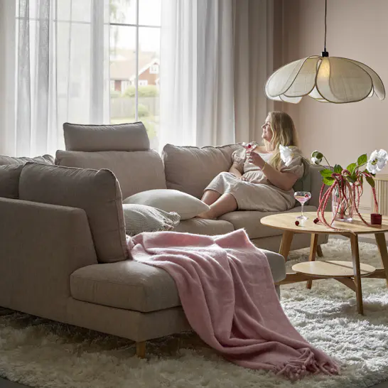En kvinna sitter i en beige soffa med divan och tittar ut genom ett fönster. På soffan ligger en rosa pläd.