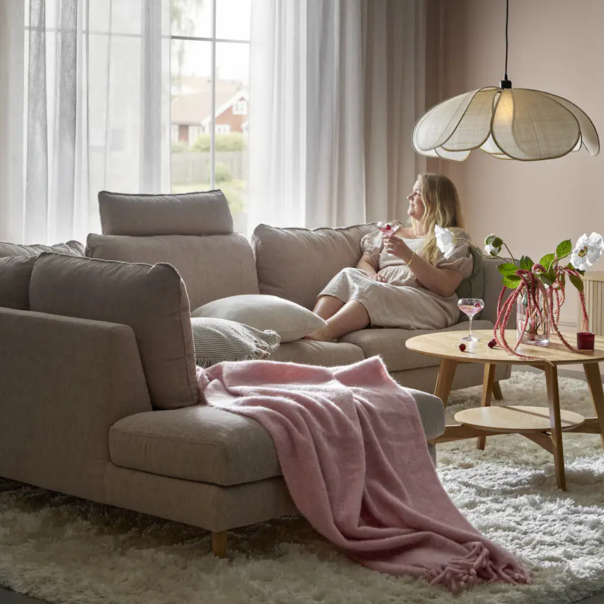 En kvinna sitter i en beige soffa med divan och tittar ut genom ett fönster. På soffan ligger en rosa pläd.