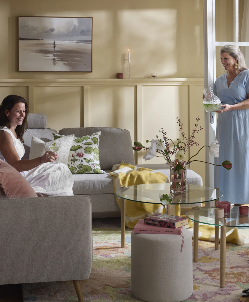 två kvinnor i ett vardagsrum varav den ena sitter i en grå hörnsoffa och den andra står i dörröppningen till trädgården