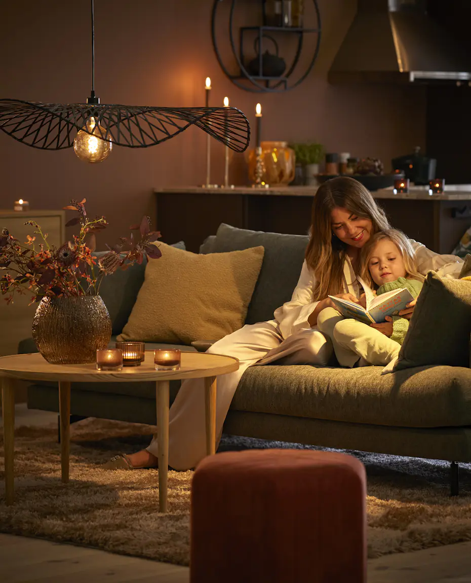 I en soffa i ett mysigt belyst vardagsrum sitter en kvinna och läser med ett barn i famnen.