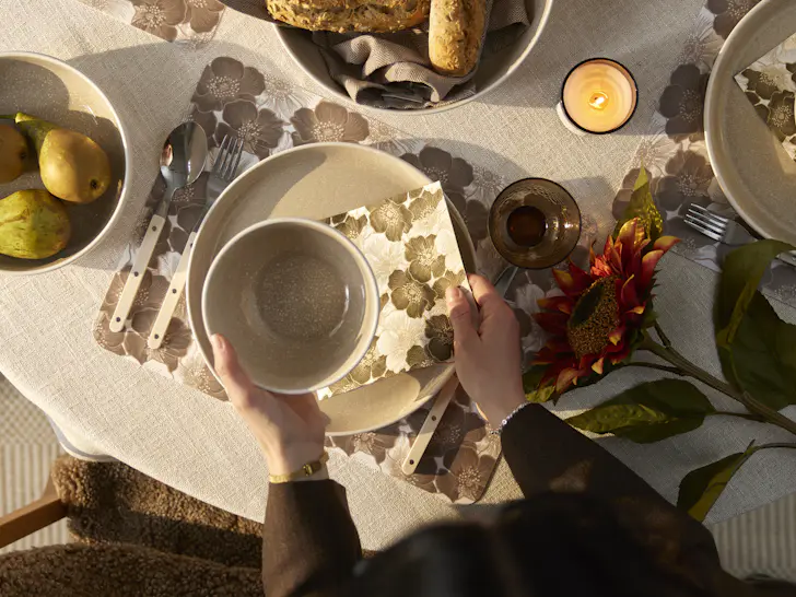 Uppduktat frukostbord med beige servis, beige duk på bord och solros i vas. Färska frallor och päron på fat
