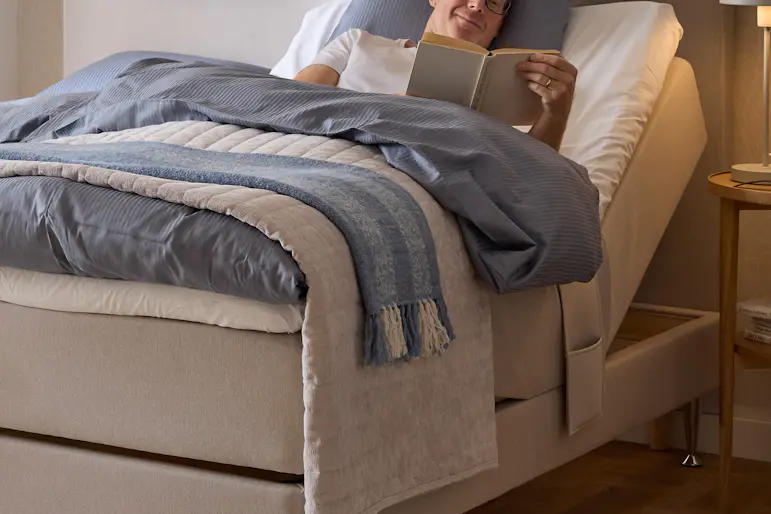 En man ligger i en ställbar beige säng och läser en bok