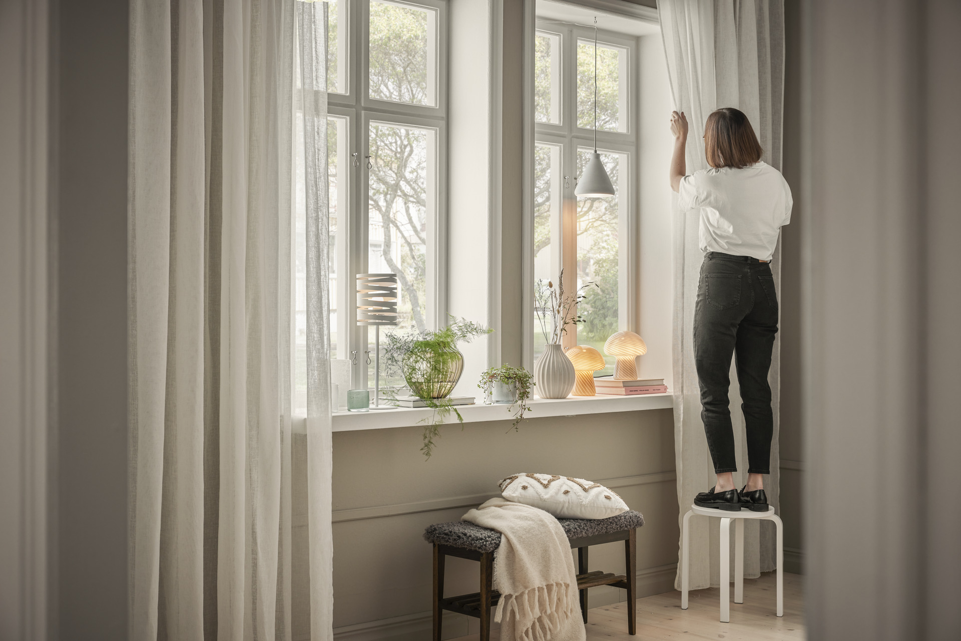 I ett fönster står en kvinna på en pall och hänger upp gardiner