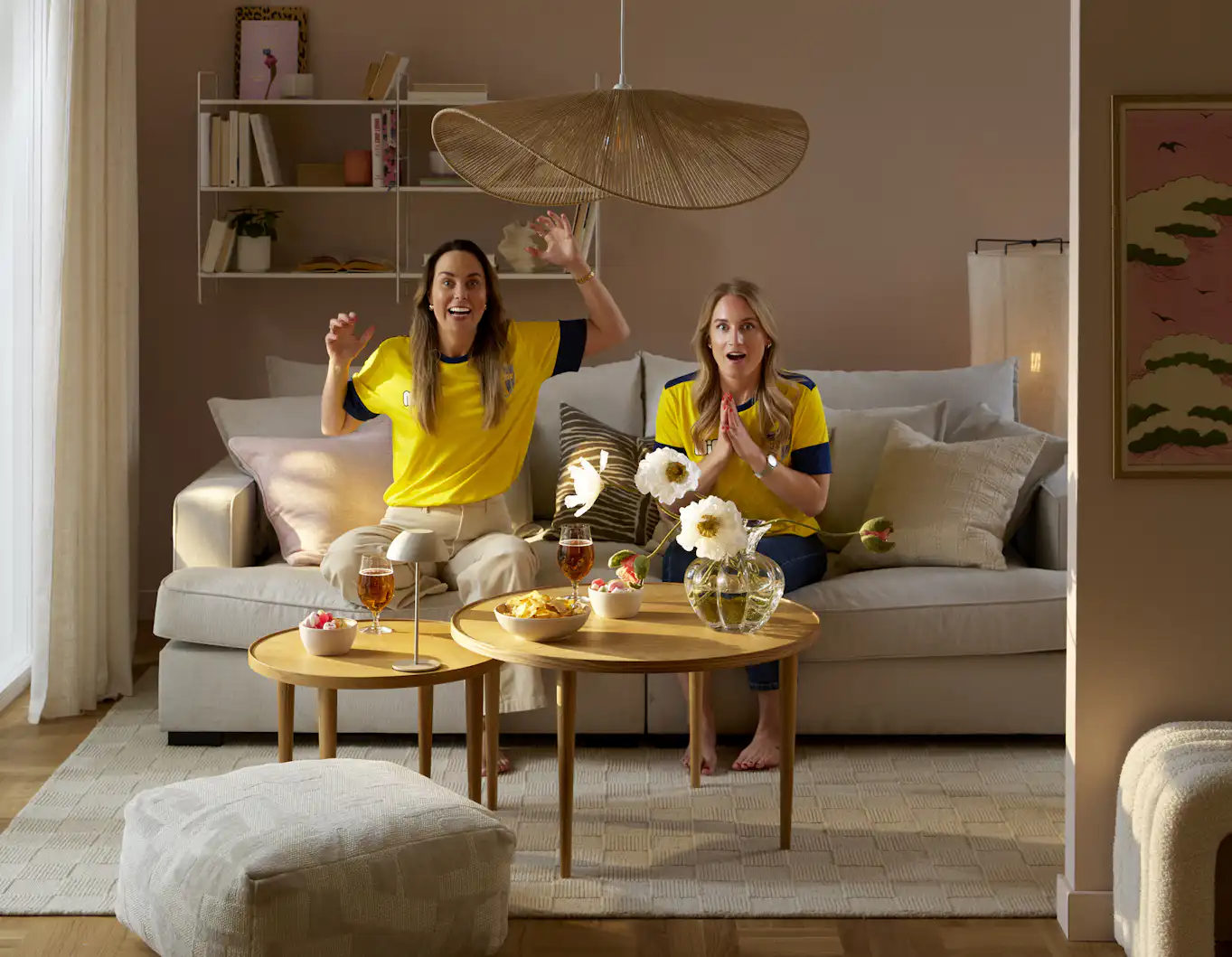 Två tjejer i gula landslagströjor sitter i soffa vända mot tv och ser ut som att något spännande händer på tv.