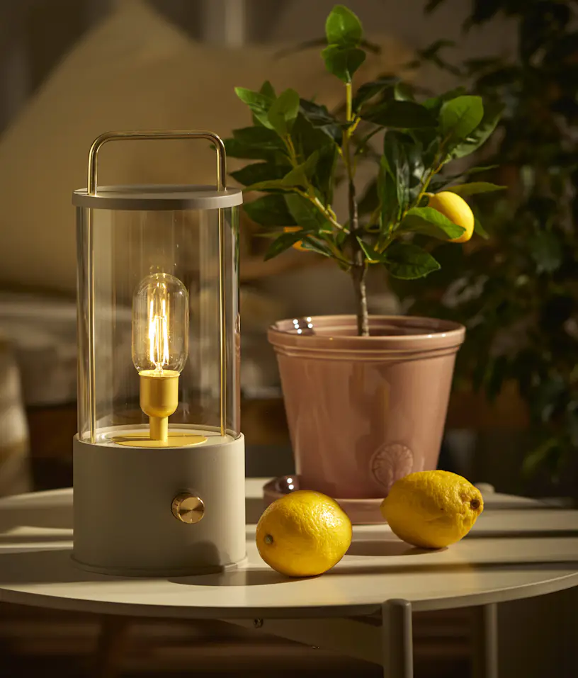 En portabel beige bordslampa står på ett utesoffbord intill en kruka med en citrusväxt.