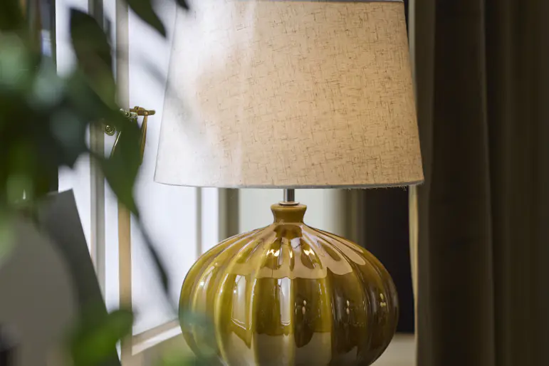 Bordslampa med fot i mörkgrön keramik och ljus lampskärm står i ett fönster.