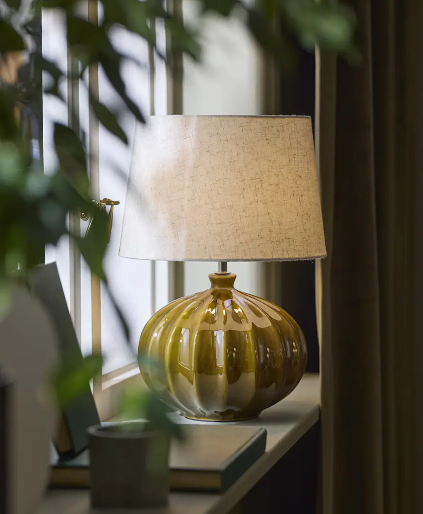 Bordslampa med fot i mörkgrön keramik och ljus lampskärm står i ett fönster.