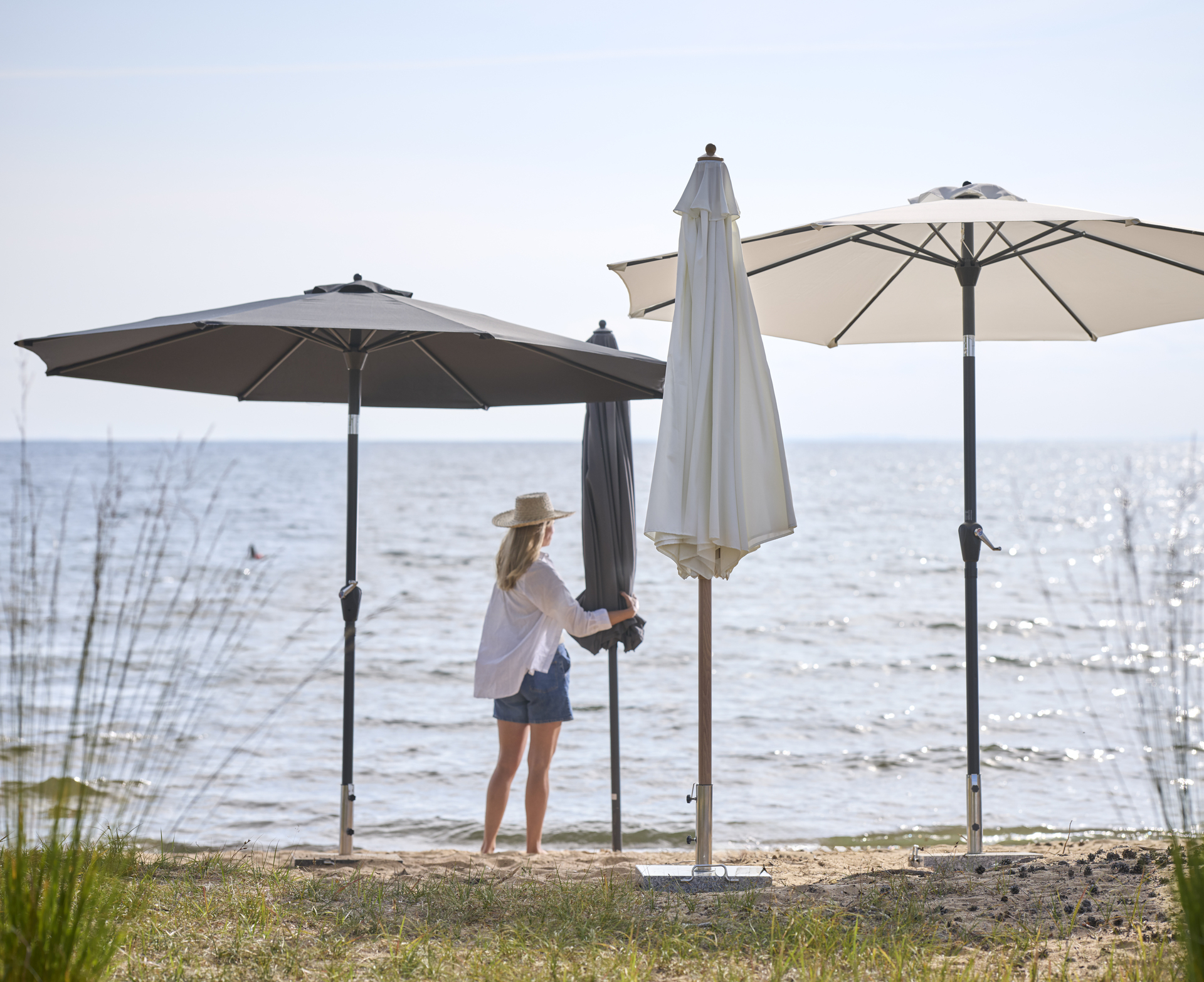 en kvinna i vit skjorta och shorts står på stranden bland flera parasoll i olika färger