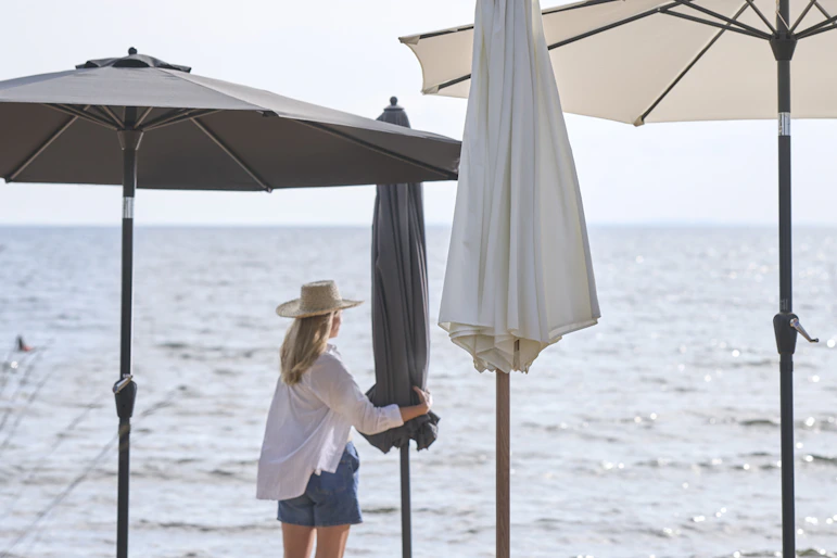 en kvinna i vit skjorta och shorts står på stranden bland flera parasoll i olika färger