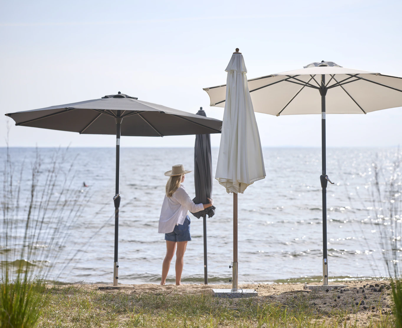 en kvinna i vit skjorta och shorts står på stranden bland flera parasoll i olika färger