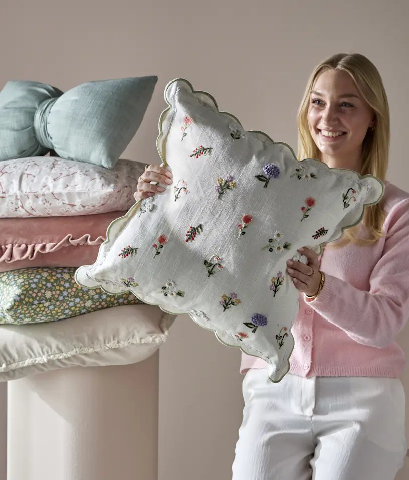 kvinna håller upp en vit kudde med broderade blommor. Bredvid henne ligger prydnadskuddar i flera olika färger staplade på en piedestal.