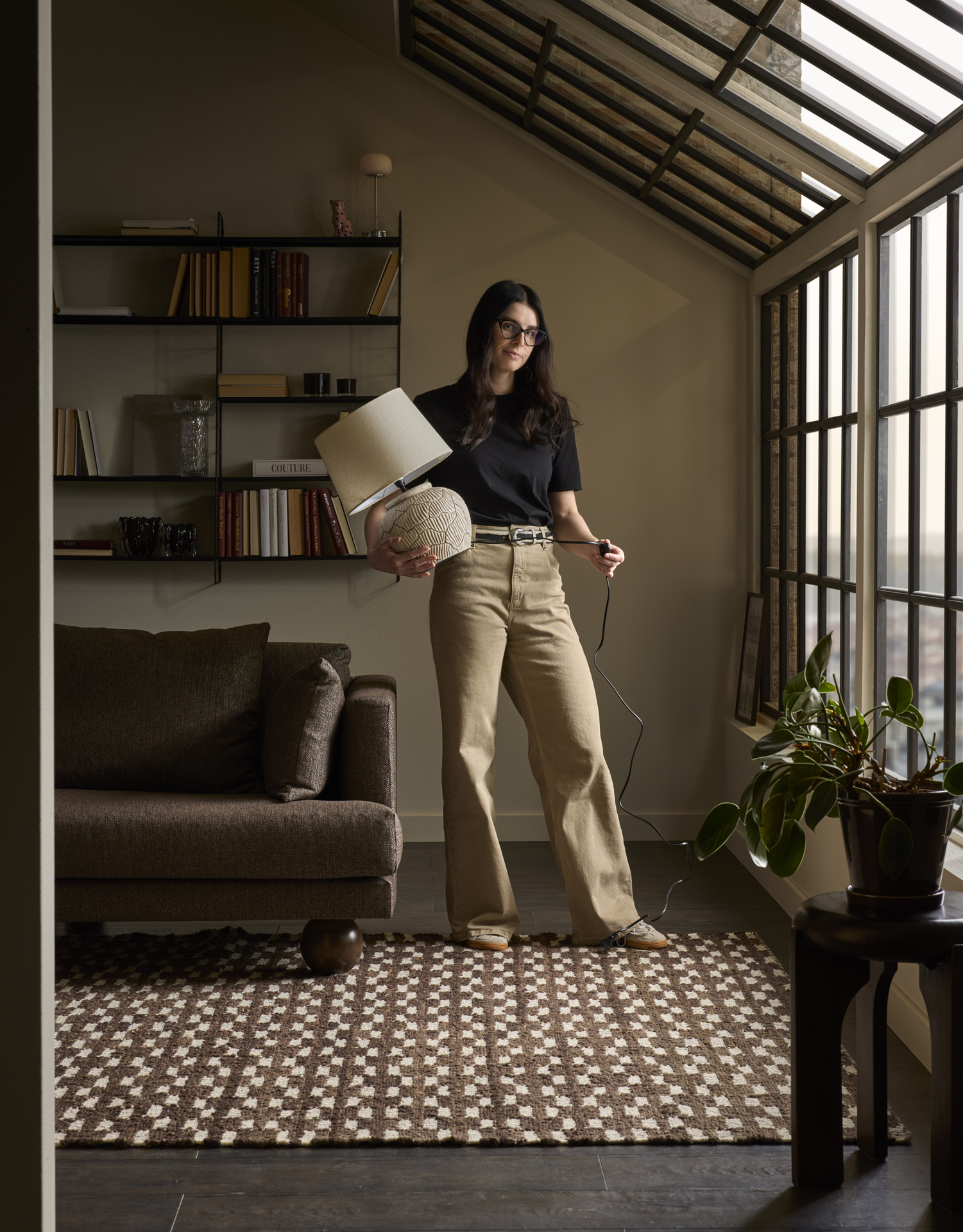 Joanna Vukota Carlsson, stylist på Mio, står i ett rum med mustiga, varma toner och håller i en bordslampa