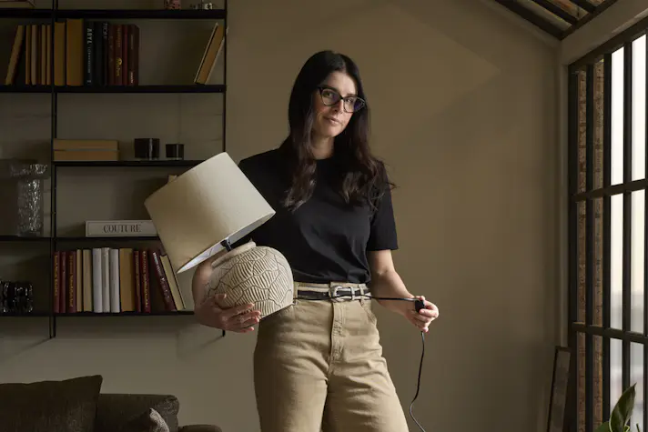 Joanna Vukota Carlsson, stylist på Mio, står i ett rum med mustiga, varma toner och håller i en bordslampa