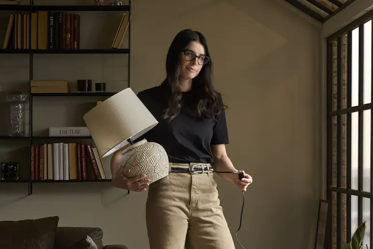 Joanna Vukota Carlsson, stylist på Mio, står i ett rum med mustiga, varma toner och håller i en bordslampa