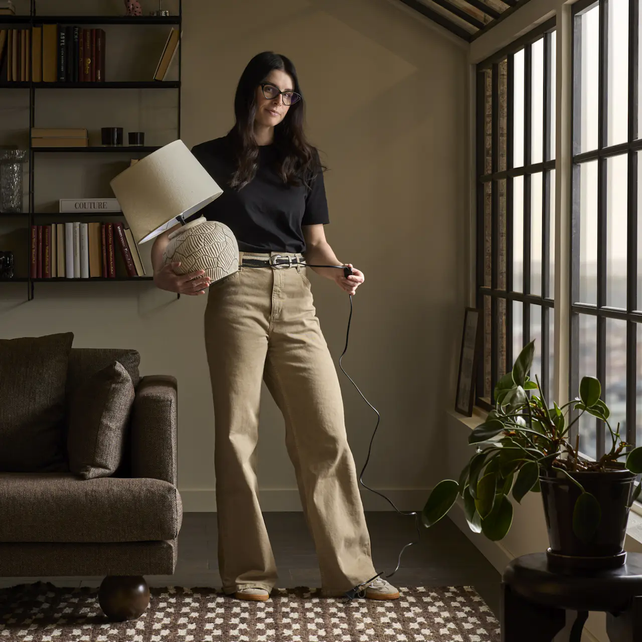 Joanna Vukota Carlsson, stylist på Mio, står i ett rum med mustiga, varma toner och håller i en bordslampa