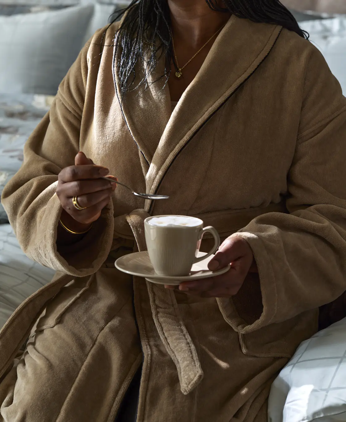En kvinna iklädd en beige Mayfair badrock håller i en kopp kaffe och en tesked.