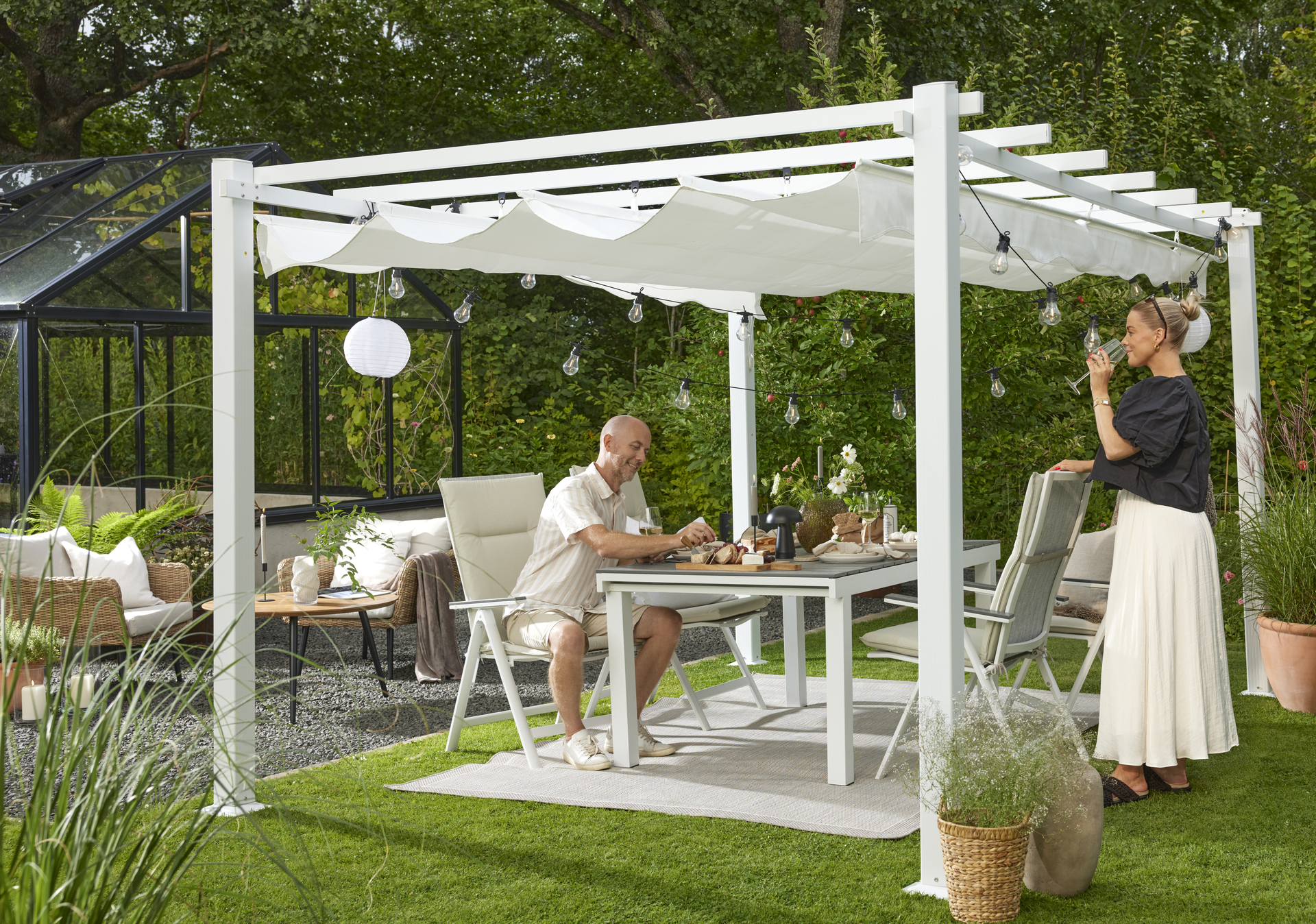 En man och en kvinna sitter och står vid en utegrupp under en pergola i en trädgård
