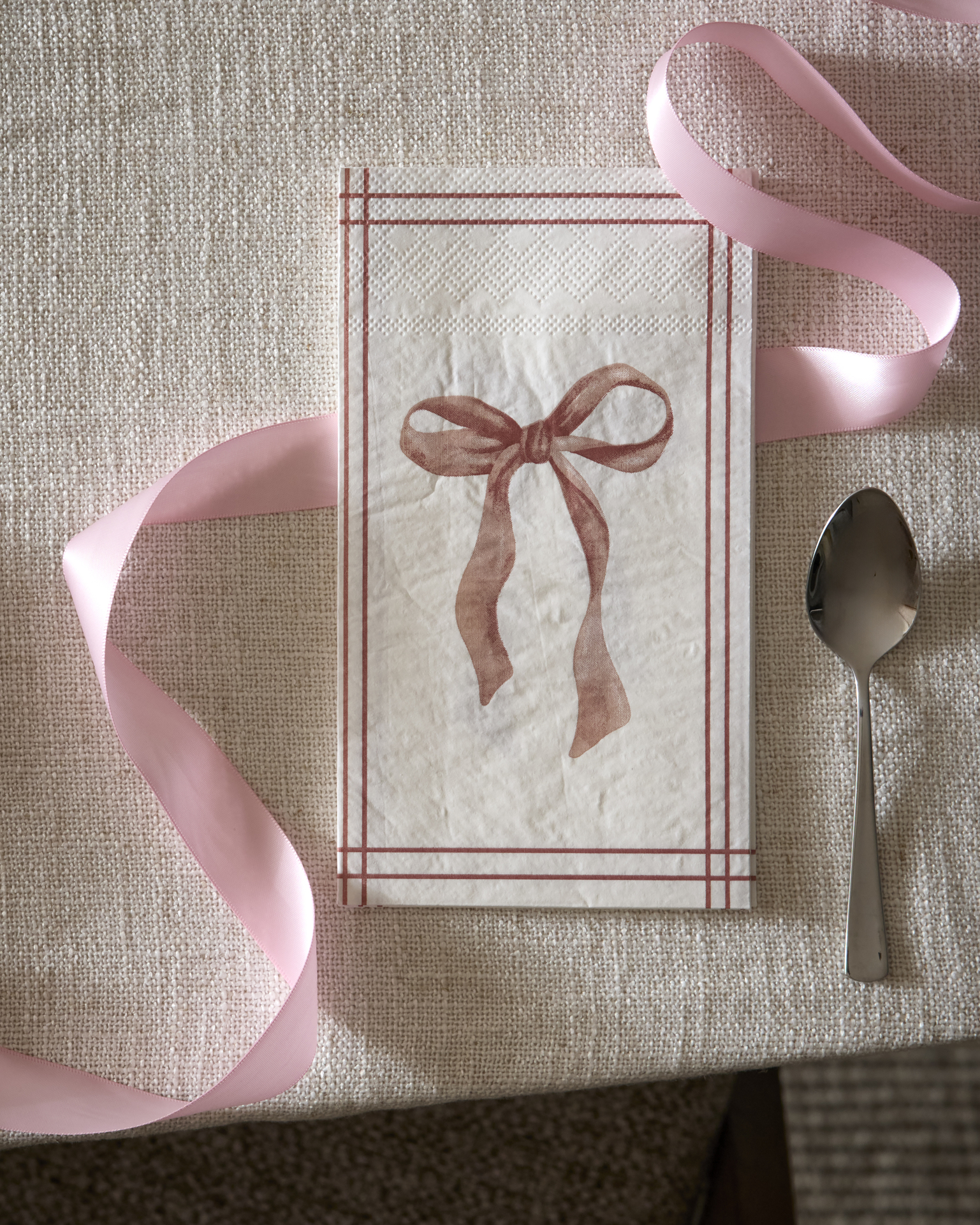 På ett bord med beige duk ligger en servett med en rosa rosett som motiv, samt ett rosa band och en tesked.